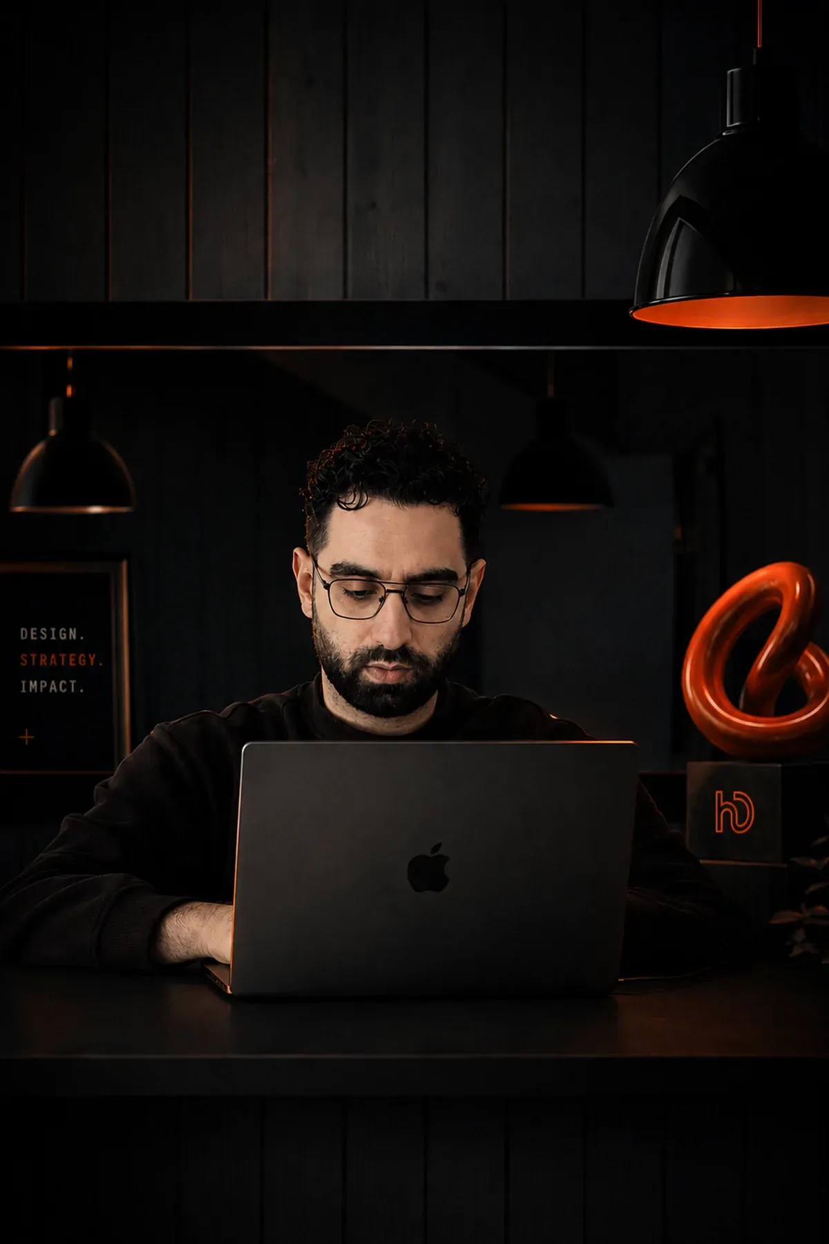 Hussein Deeb at his desk in the dark studio, working at a laptop under pendant lamps.