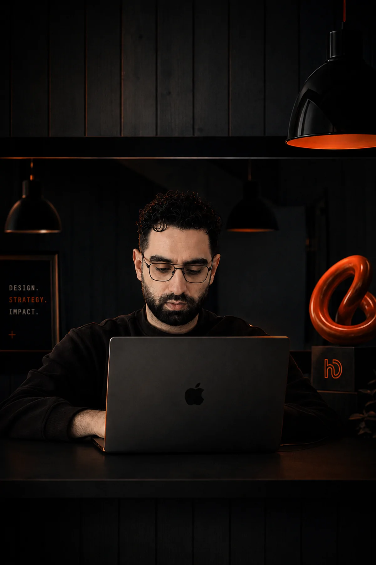 Hussein Deeb at his desk in the dark studio, working at a laptop under pendant lamps.